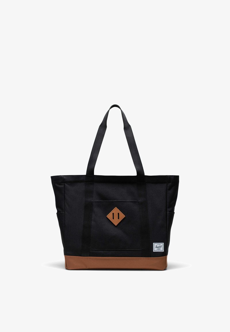 Bolsa de tela negra con acento en la parte inferior marrón, que cuenta con dos asas resistentes y un parche de cuero en forma de diamante en la parte frontal.