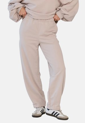 Osoba ubrana w beżowe spodnie dresowe i pasującą, oversize'ową bluzę, stojąca z rękami w kieszeniach, nosząca białe i czarne sneakersy.
