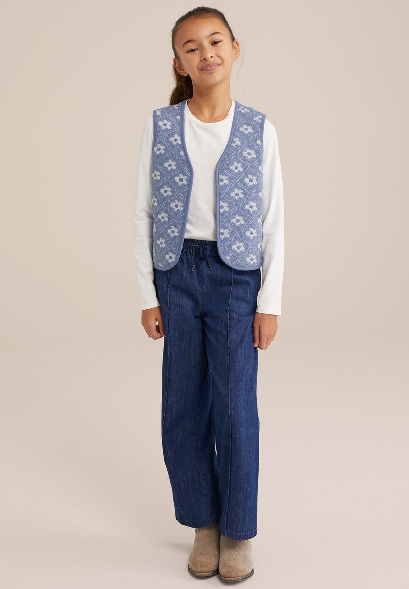 Blau gemusterte Weste mit weißem Blumenmuster über einem weißen Langarmshirt, kombiniert mit dunkelblauen weiten Hosen und beigefarbenen Stiefeletten.
