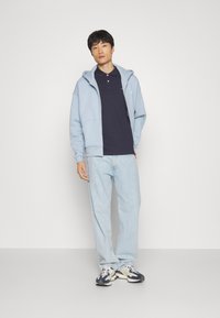 Lys blå hettegenser med glidelås over en marineblå polo, kombinert med lyse denimjeans og mørke sneakers, stående foran en ensfarget bakgrunn.