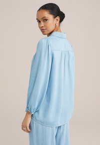 Hellblaues Denimhemd mit lockerem Schnitt, langen Ärmeln und Raffung an den Manschetten. Verfügt über eine Rückenpasse und einen abgerundeten Saum.