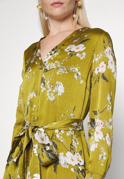 Vestido floral verde com um tecido texturizado, apresentando um decote em V, botões e um accentuador de cintura amarrado. Padrões florais em branco e laranja.