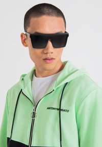 Occhiali da sole neri e oversize con forma angolare, abbinati a una felpa verde chiaro con zip e dettagli neri e una cerniera argentata.