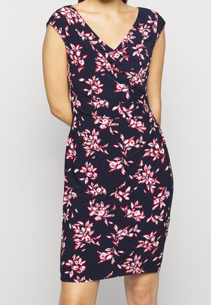 Woman wearing a sleeveless navy blue dress with red and white floral pattern and a V-neckline, standing with arms behind her back.