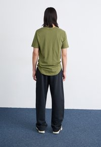 Olivgrünes Baumwoll-T-Shirt mit gerundetem Saum, kombiniert mit lockeren schwarzen Hosen. Das Outfit zeichnet sich durch ein einfaches Design und eine weiche Textur aus.