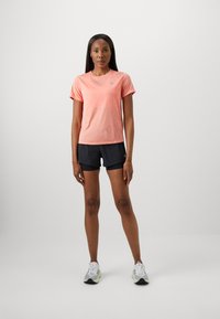 Γυναίκα με κοντομάνικο πουκάμισο κοραλί, μαύρες αθλητικές βερμούδες και λευκά αθλητικά παπούτσια, που στέκεται μπροστά σε απλό φόντο.