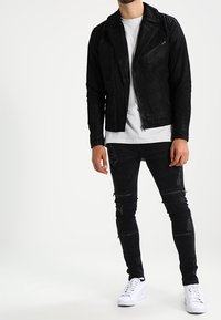 Chaqueta de cuero negra para motocicleta con diseño ajustado, llevada sobre una camiseta blanca, combinada con vaqueros pitillo negros desgastados y zapatillas blancas.