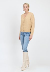 Beiger Strickcardigan mit V-Ausschnitt, Knopfverschluss und geripptem Saum, kombiniert mit hellblauen Jeans und beigen kniehohen Absatzstiefeln.