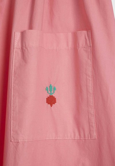 Tissu rose avec une poche rectangulaire présentant un petit radis rouge brodé avec des feuilles vertes centré sur la poche.