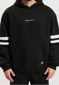 Zwarte hoodie met witte scriptlogo en witte strepen op de mouwen. Heeft een voorkantzak en geribde manchetten voor een veilige pasvorm.