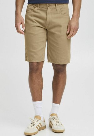 Mand iført beige knælange shorts, hvide strømper, beige og hvide sneakers, stående mod en ensartet lys baggrund.