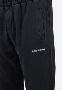 Mustad mustri püksid elastse vöökoha, küljetaskud ja valge "PEGADOR" logo paremal reiel. Pehme tekstuur ja juhuslik disain.