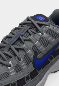Κοντινό πλάνο γκρι αθλητικού παπουτσιού Nike με μπλε λογότυπο Swoosh, μαύρη ετικέτα στη γλώσσα, παχιά γκρι κορδόνια και λεπτομέρειες από υφασμάτινο δίχτυ.