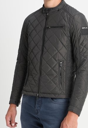 Chaqueta negra acolchada con cremallera, cuello alto y bolsillos con cremallera en el pecho y los costados, combinada con jeans azules.