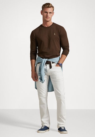 Brown long-sleeve shirt, white pants, light blue denim tied around waist, blue sneakers. Features a small logo on the shirt and a watch.