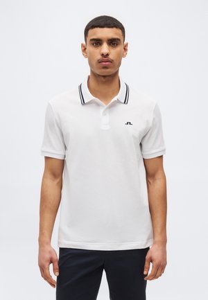 Tânăr cu păr scurt și mustață, purtând un tricou polo alb cu margini negre la guler și pantaloni închiși la culoare, stând în picioare pe un fundal alb.