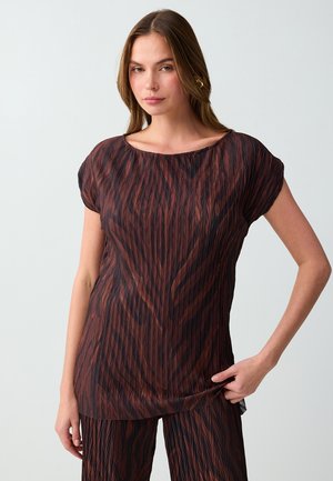 Jonge vrouw die een bruin-zwart gestructureerd kort mouw topje en bijpassende broek draagt, staande tegen een effen lichtgrijze achtergrond.