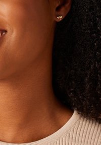 Goldfarbene Bienen-Ohrringe mit kleinen klaren Strasssteinen, die eine glatte Oberfläche und filigrane Details aufweisen, getragen vor einem strukturierten Hauthintergrund.