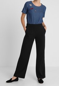 Camiseta de denim de manga corta con tres parches decorativos; combinada con pantalones anchos negros y zapatos negros tipo slip-on.
