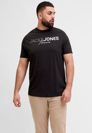 Mand med skæg iført sort Jack & Jones Premium t-shirt og beige bukser, der kigger til venstre mod en ensartet lys baggrund.
