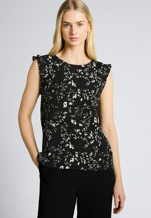 Femme blonde aux cheveux lisses portant un haut noir sans manches à motifs floraux et un pantalon noir sur un fond gris clair uni.