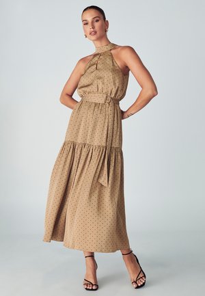 Tan maxi dress with polka dots, halter neckline, cinched waist with a belt, and tiered skirt; worn with strappy black heels.