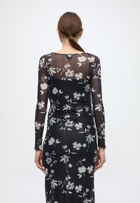 Μακρυμάνικο, διάφανο μαύρο φόρεμα διακοσμημένο με φωτεινά λουλουδένια σχέδια. Εφαρμοστή σιλουέτα, με ανοιχτή πλάτη και λεπτομέρειες από βολάν στους μανσέτες.