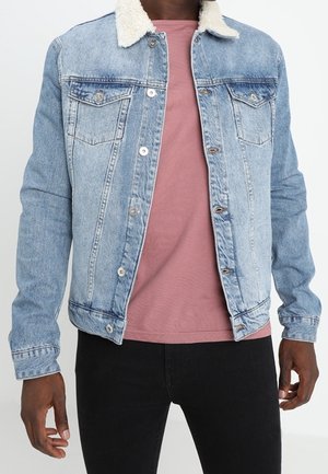 Mężczyzna ubrany w jasnoniebieską kurtkę dżinsową z białym kołnierzem typu sherpa, różową koszulkę w kolorze pudrowego różu oraz czarne spodnie, stojący na białym tle.