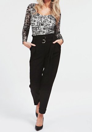 Mujer vestida con pantalones negros de talle alto y cinturón, y una blusa de manga larga con un patrón en blanco y negro, con las manos en los bolsillos y tacones negros.