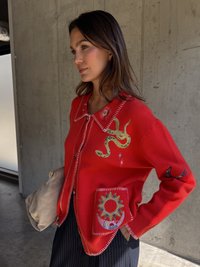 Piros kardigán kígyó és égi hímezéssel, kontrasztos fehér varrással, valamint egy nap mintázatú zsebbel. Texturált anyagból készült.