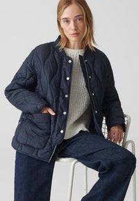Giacca di pelle blu navy trapuntata con bottoni a pressione argentati, scollatura tonda e tasche laterali, abbinata a un maglione lavorato a maglia color crema e jeans blu scuro.