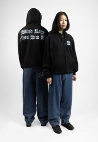 Zwarte zet-rits hoodie met blauwe grafische tekst op de achter- en voorkant, gecombineerd met losse blauwe denim jeans met een spinnenwebontwerp op de zak.