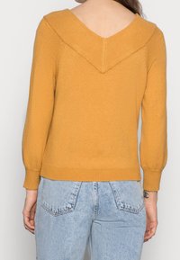 Pull en maille jaune moutarde avec un large col en V, col côtelé et manches ballon, associé à un jean taille haute bleu clair.