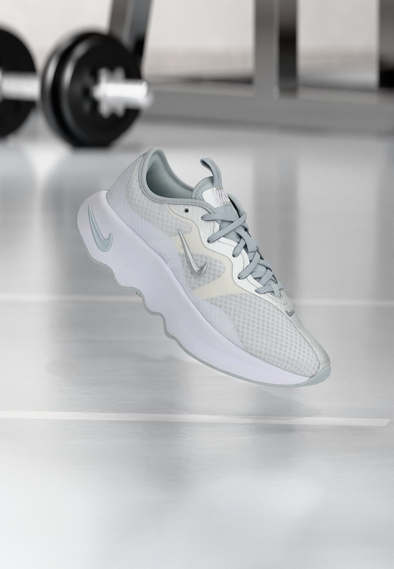 Γκρι αθλητικό παπούτσι Nike με πλεκτό σχέδιο και παχιά σόλα, αιωρούμενο πάνω από μια αντανακλαστική επιφάνεια με θαμπά βάρη γυμναστηρίου στο βάθος.