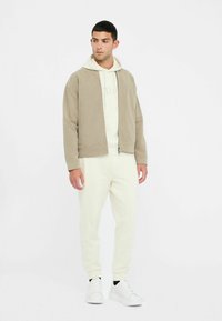 Brun kordfløjl zip-jakke over en creme hoodie, parret med creme joggers og hvide sneakers. Sættet har enkle linjer og en afslappet pasform.