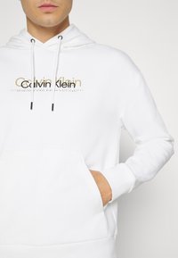 Felpa con cappuccio bianca, con tasca frontale e cordini, decorata con il logo oro e nero di Calvin Klein nella zona del petto.
