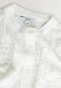 Balta mežģīņu virsjaka ar apaļu izgriezumu, pogu aizdari un sarežģītiem ziedu rakstiem. Izgatavota no viegla auduma ar mīkstu tekstūru.