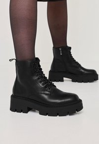 Bottines montantes en cuir noir à lacets avec semelles épaisses, portées avec des collants noirs transparents sur fond clair.