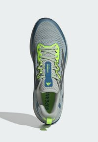 Harmaa urheilujalkine, jossa on verkkomateriaali ja vihreät yksityiskohdat, pehmustettu nilkkapanta ja paksu pohja. Sisältää heijastavia yksityiskohtia ja logon.