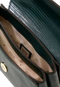 Intérieur texturé vert d'un sac à main avec deux compartiments, une fermeture à bouton doré et une étiquette logo GUESS sur une doublure beige.