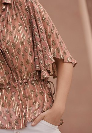 Blouse rose avec motifs floraux métalliques dorés, manches volantées, taille à cordon, et tissu léger texturé.