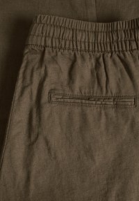 Braune Stoffshorts mit einem elastischen Bund und einer flachen Gesäßtasche mit gesticktem Rand. Die Stoffstruktur wirkt weich und leicht zerknittert.