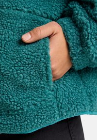 Teal fleecestijljas met een gestructureerd, pluizig oppervlak; heeft een zijzak met een hand die gedeeltelijk zichtbaar is en de stof vasthoudt.