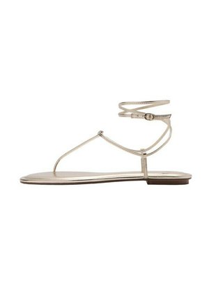 Sandale plate en métal doré avec fines lanières, boucle à la cheville et design à bride en T sur fond blanc.