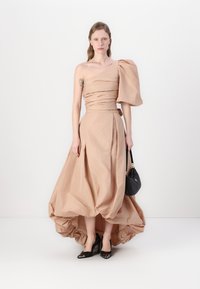 A nude, one-shoulder dress featuring a structured puff sleeve, draped detailing, and a high-low hemline, paired with black pointed heels and a handbag.