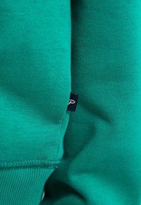Turquoise katoenen sweatshirt met ribbelstructuur en zichtbare stiksels, met een klein donkerblauw label van stof dat in de naad is genaaid.