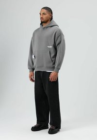 Grijze oversized hoodie met een voorzak, wit logodetail, zwarte losse jeans en zwarte veter schoenen. Soepele stoftextuur.
