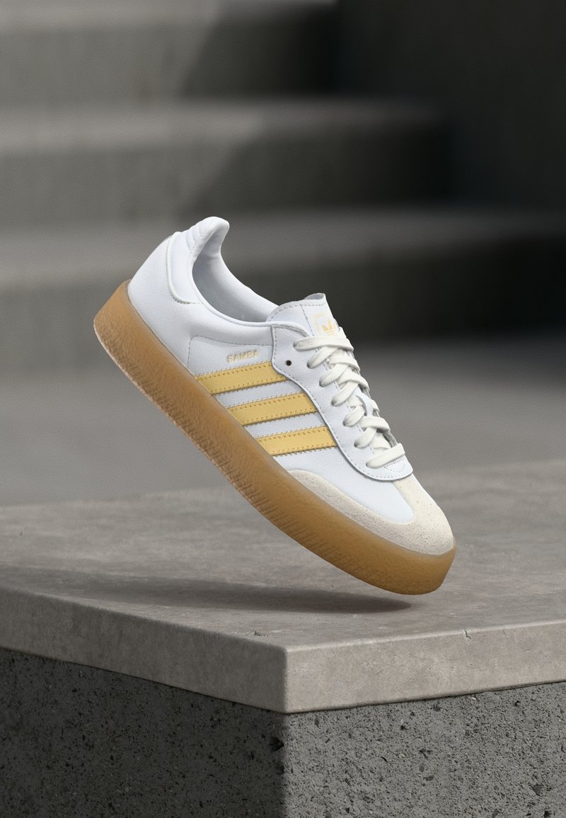 Biały skórzany sneaker z żółtymi akcentami, trzema paskami i gumową podeszwą. Posiada zaokrąglony nosek i teksturowany język.