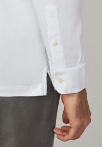 Weißes Hemd mit Knopfleiste, das einen spitzen Manschette mit zwei Knöpfen, eine glatte Stofftextur und einen seitlichen Schlitz am Saum aufweist. Getragen mit grauen Hosen.