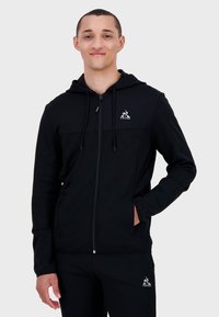 Felpa nera con zip realizzata in morbido tessuto, con tasca a marsupio, coulisse regolabile e logo sul petto.